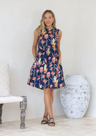 Woman wearing a pink and green floral print on a blue base sleeveless dress with hands in side pockets
