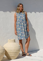 Woman standing in a relaxed fit above knee length dusty blue floral cotton dress with pockets, sleeveless style and high round neckline 