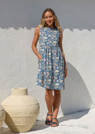 Woman in a above knee length 100% cotton floral dress standing against a white wall with a large beige pot nearby