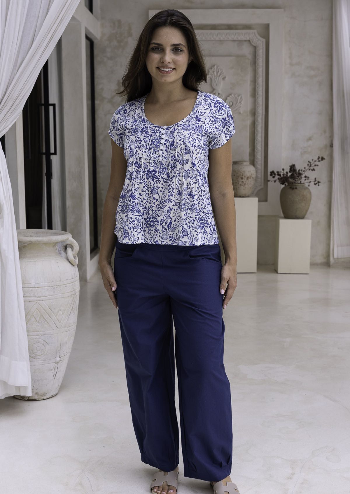 Woman wearing a white based blue floral print cotton top with dark pants.