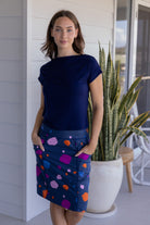 A woman standing in a modern room, wearing a knee-length skirt with a multicoloured spot print, with her hands in pocket