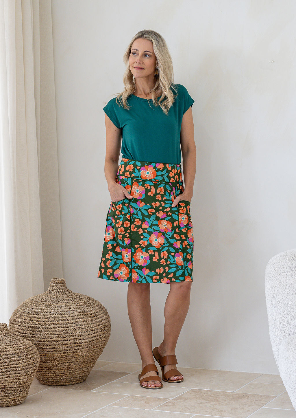 Woman standing with both hand in pockets wears olive green based floral print cotton above knee length skirt piped detailing.