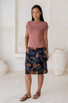 Woman standing with hand in pocket wears black cotton above knee length skirt with colorful botanical print and piped detailing.
