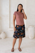 Woman wearing a dusty pink top and floral  cotton skirt in black with A-line shape, piped detailing and pockets.