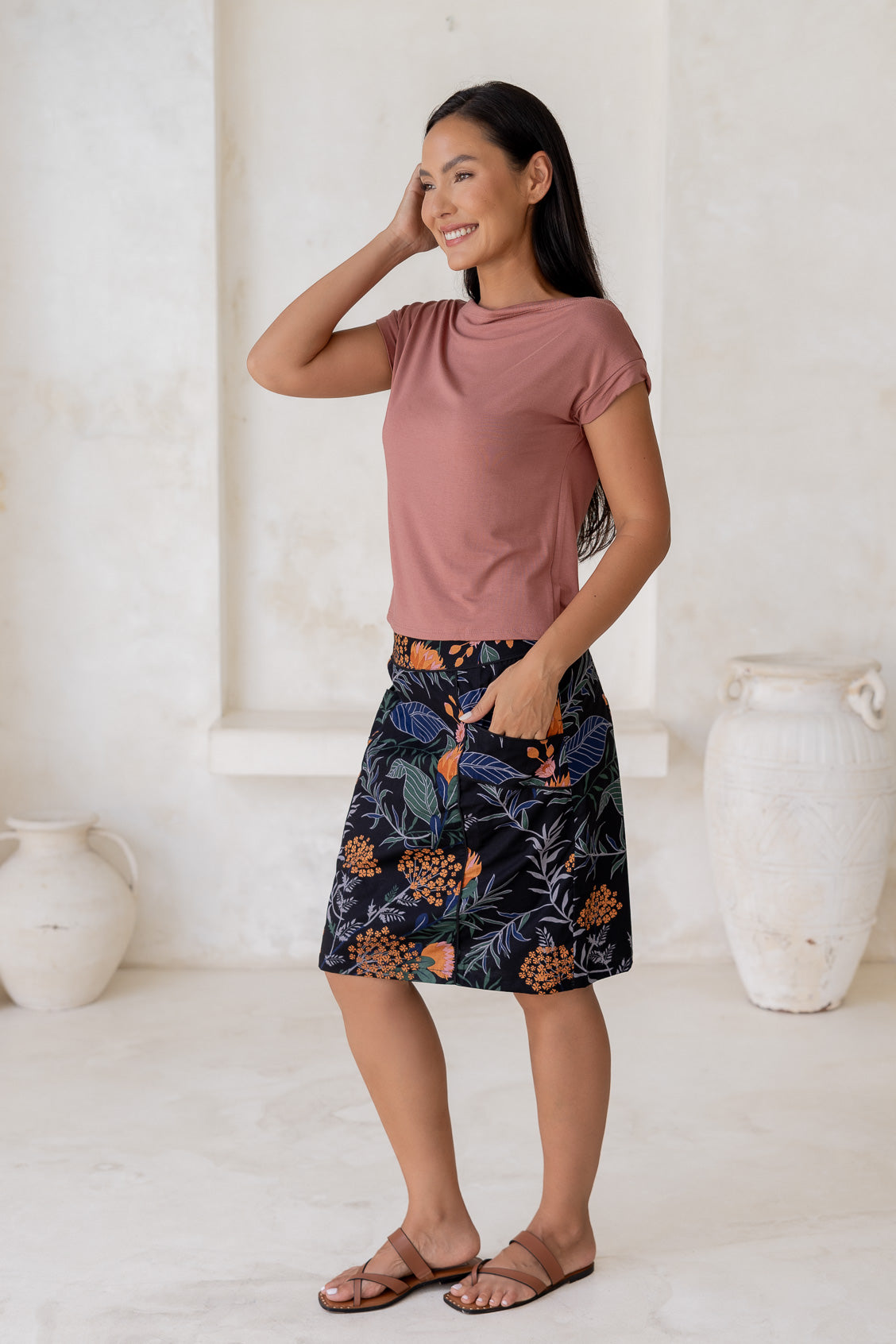 Woman posing slightly sideways wears 100% cotton A-line skirt in black based botanical print, paired with dusty rose basic top.