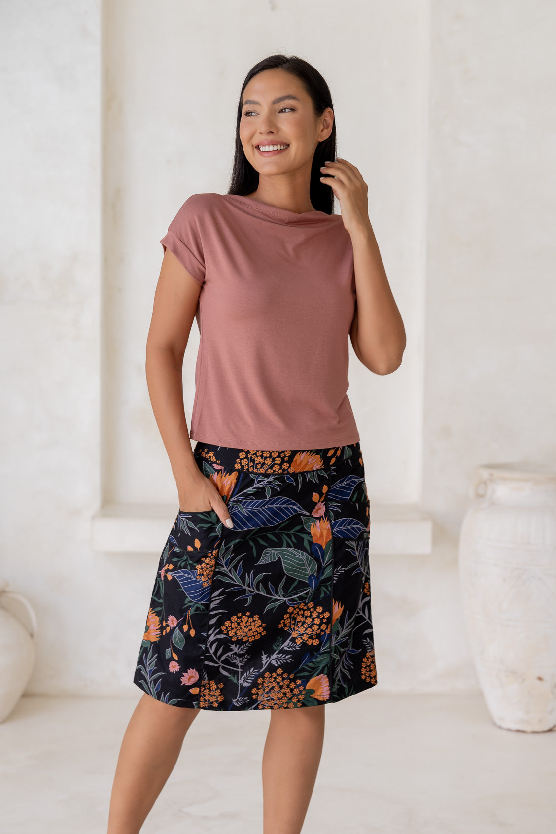 Woman stands in 100% cotton above knee length A-line skirt with piped detailing, pockets and a side zipper.
