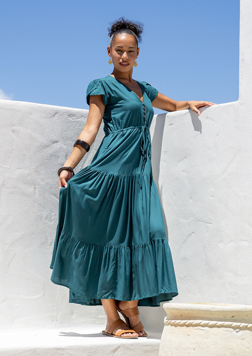 Woman in a teal maxi dress standing against a white wall with blue sky.