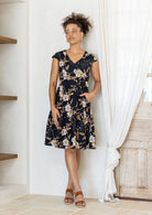 Model with one hand in pocket wears 100% cotton dress with crossover bodice, cap sleeves and fuller skirt in black based floral print.
