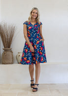 Model with one hand in pocket wears 100% cotton dress with crossover bodice, cap sleeves and fuller skirt in royal blue based floral print.