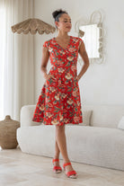 Model with both the hands in pocket wears 100% cotton dress with crossover bodice, cap sleeves and fuller skirt in red based floral print.