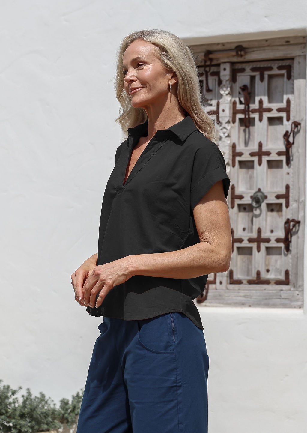 Woman wearing a black cotton collared short sleeved shirt and blue pants standing in front of a white wall