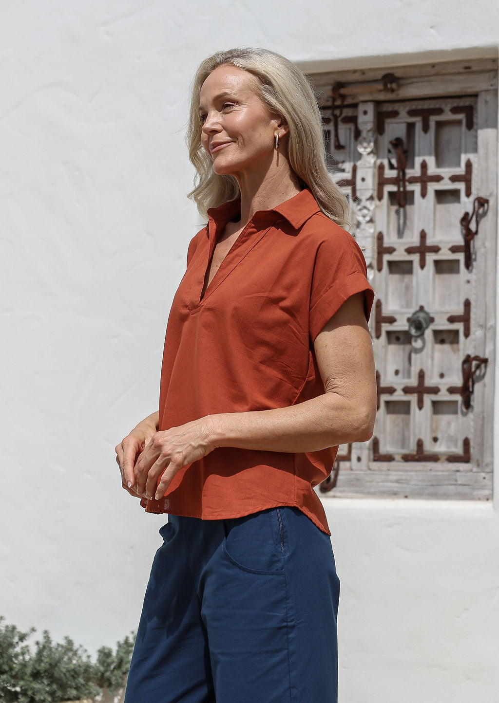 Woman standing slightly sideways wears 100% cotton shirt style top in brick orange shade, paired with sea blue cotton pants