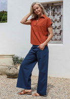 Woman wearing relaxed fit collared top in rust orange with short sleeves and V neckline, paired with blue wide leg cotton pants  