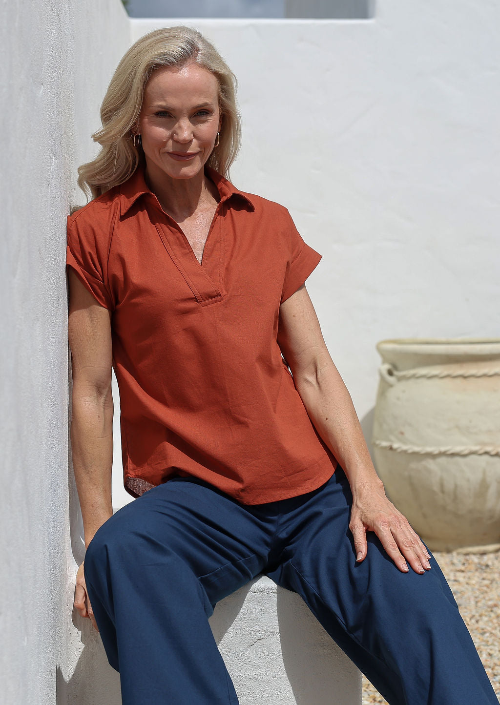 Woman wearing a rust-colored loose cut shirt style cotton top and blue pants leaning against a white wall with a decorative pot.