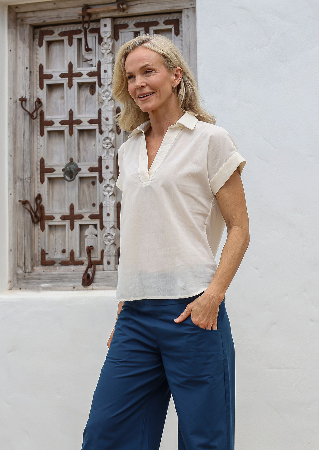Woman standing with hands in pants pockets wears lightweight 100% cotton loose fit top with collar and short sleeves, paired with sea blue cotton pants