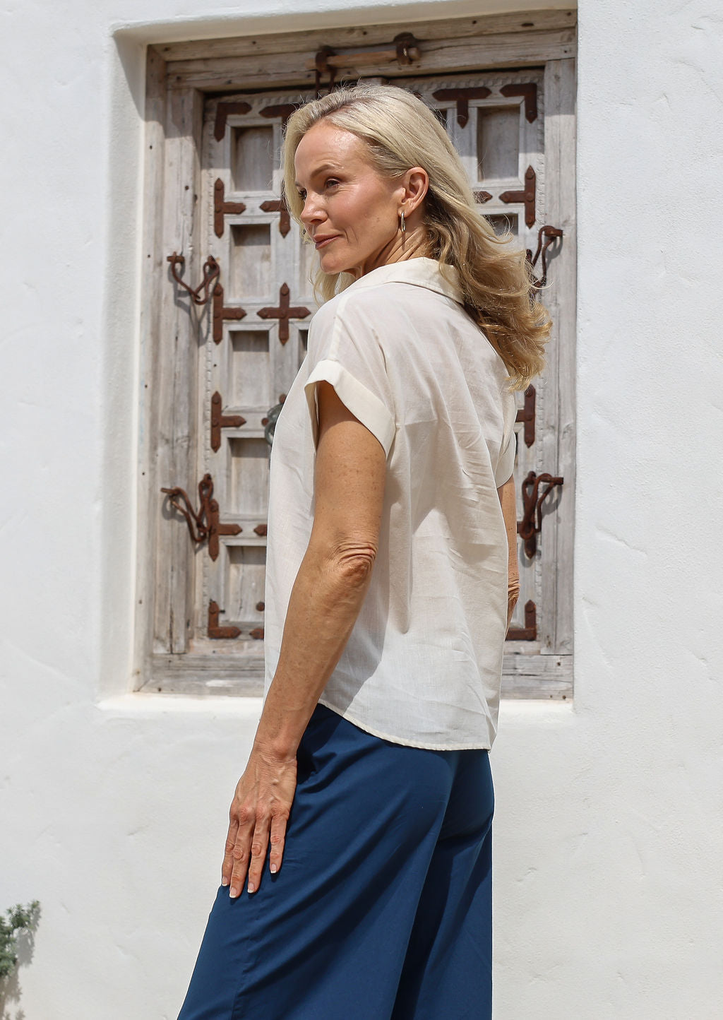 Woman wearing a white shirt style cotton blouse and blue pants standing in front of a white wall with a decorative window