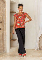 A woman poses in a cap-sleeved cotton tailored top with vibrant red backdrop accented by elegant multicoloured floral vines.