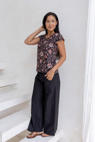 Woman posing in cap sleeved cotton top in dark brown based indian floral print, paired with wide leg black cotton pants.
