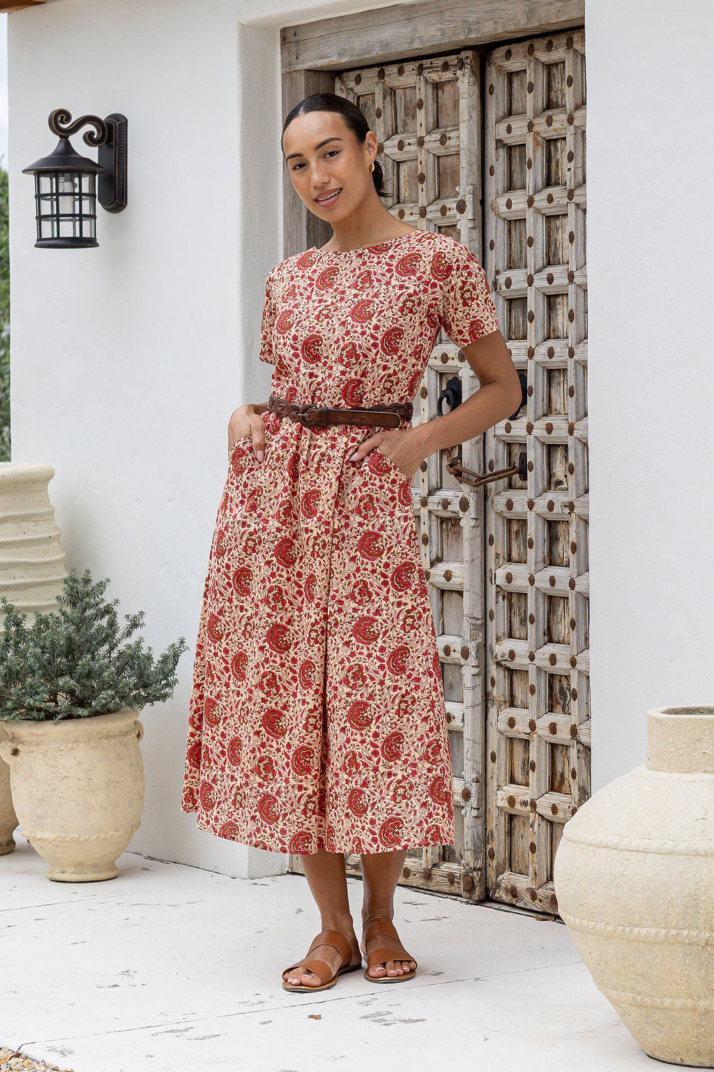 Woman poses with both hand in pockets of short sleeved midlength cotton dress with boat neckline and cream based warm floral print.