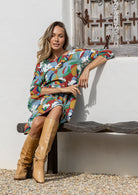 Woman in a colorful dress and tan boots sitting on a bench against a white wall.