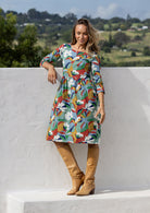 Woman in a colorful floral dress and brown boots standing against a white wall with a scenic background.