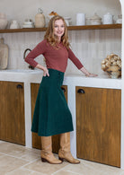 Woman standing sideways wearing a terracotta top and green cotton corduroy A-line midi length skirt with belt loops