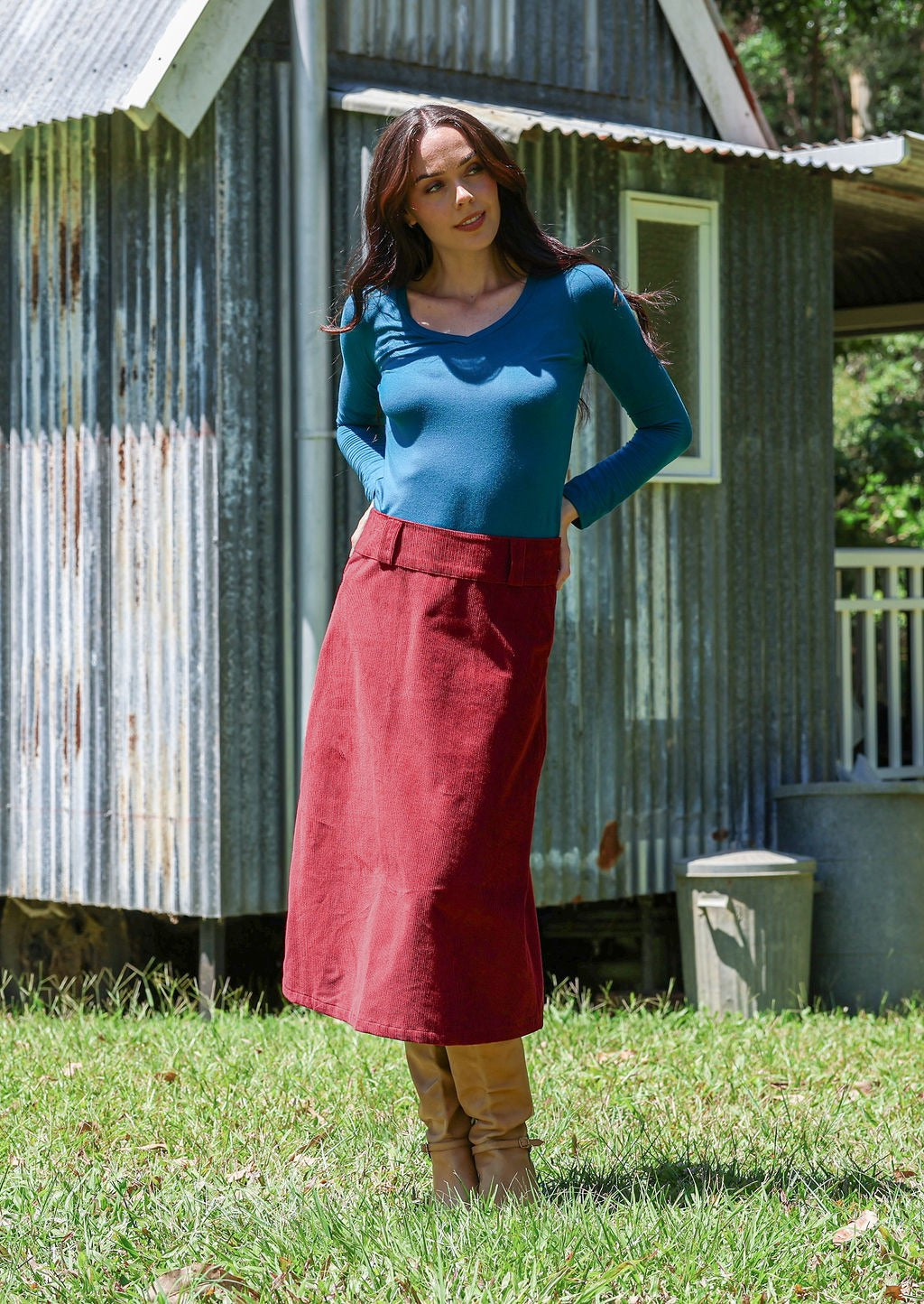 Woman wearing a cabernet red cotton corduroy A-line midi length skirt with belt loops, paired with a teal top and tan boots