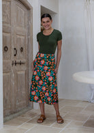 Woman stands wearing a basic top and olive green based floral print, button through cotton skirt with box pleats and belt loops 