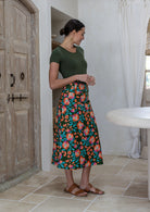 Woman stands sideways wearing a knee length, box pleated cotton skirt with button front, belt loops and pockets.