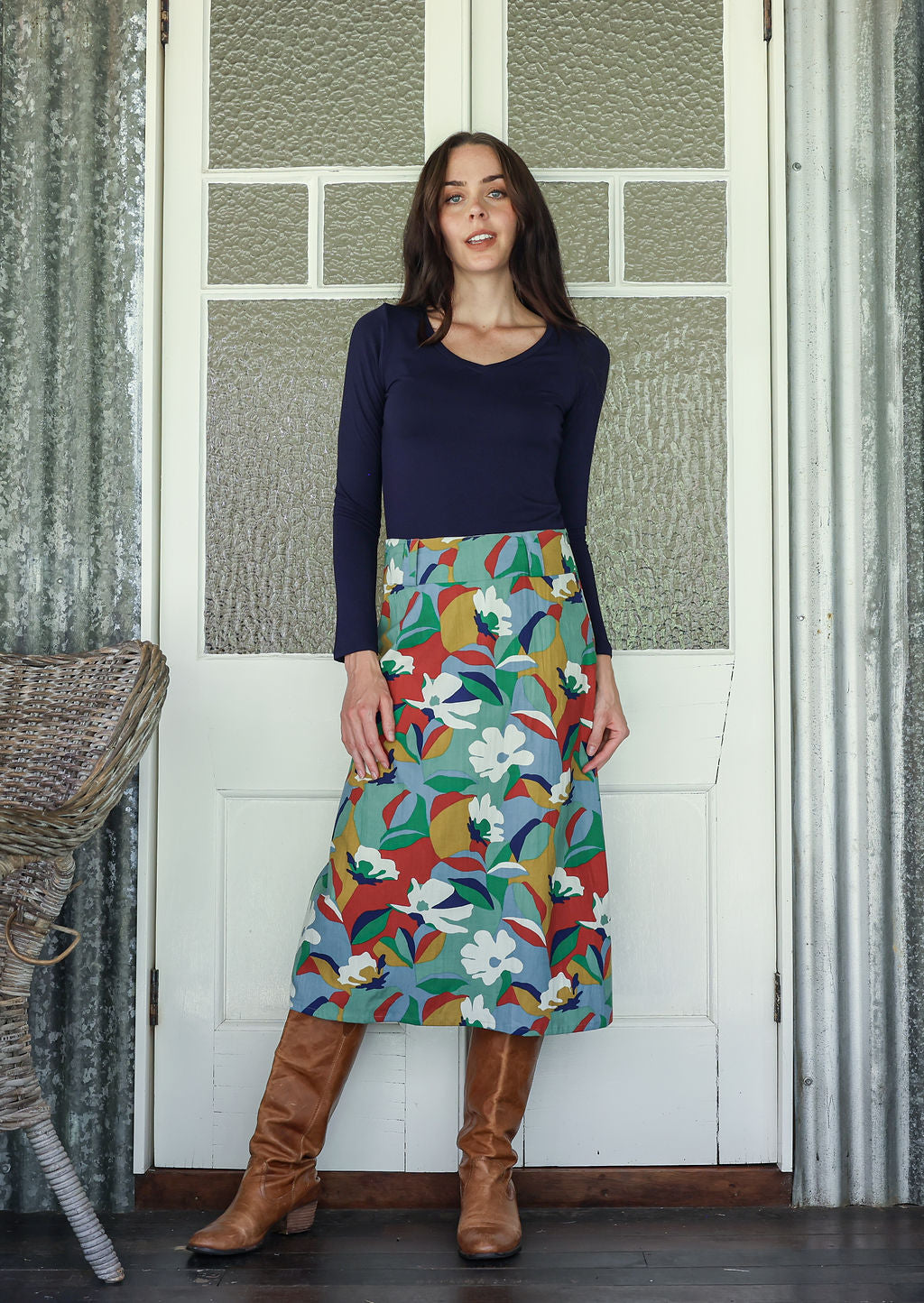 Woman wearing a colorful floral skirt and brown boots standing in front of a white door.