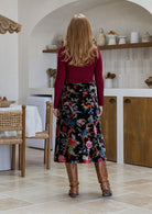 Woman standing backwards in a black based multi-coloured florals cotton velvet skirt paired with a maroon long sleeve top and tan boots
