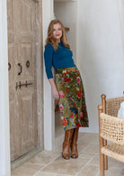 Woman standing in a moss green based multi-coloured florals cotton velvet skirt paired with a blue long sleeve top and tan boots