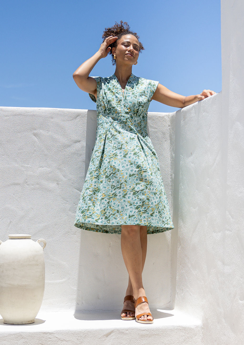 Woman wearing sage green based floral cotton dress with cap sleeves, box pleats, side zip and V neckline