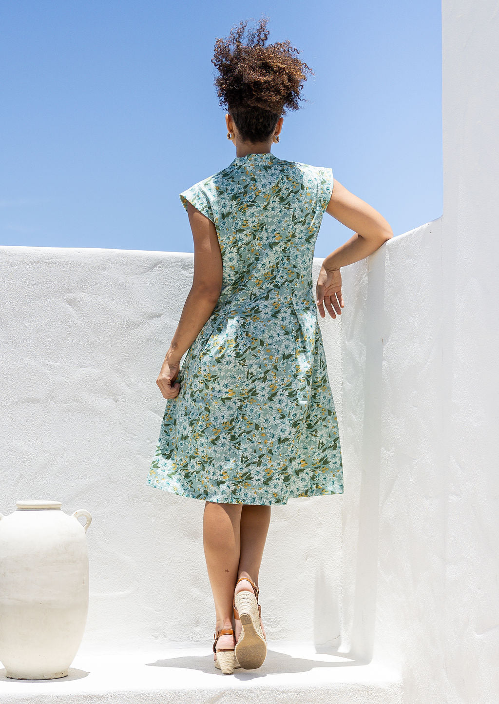 Woman stands backwards in a floral cotton dress against white wall and a blue sky