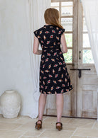 Woman stands backwards wearing 100% cotton retro style knee length dress with box pleats, cap sleeves and V neckline.