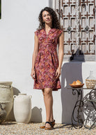 Model wearing a knee length warm toned floral cotton dress with cinched waist, box pleats and cap sleeves 