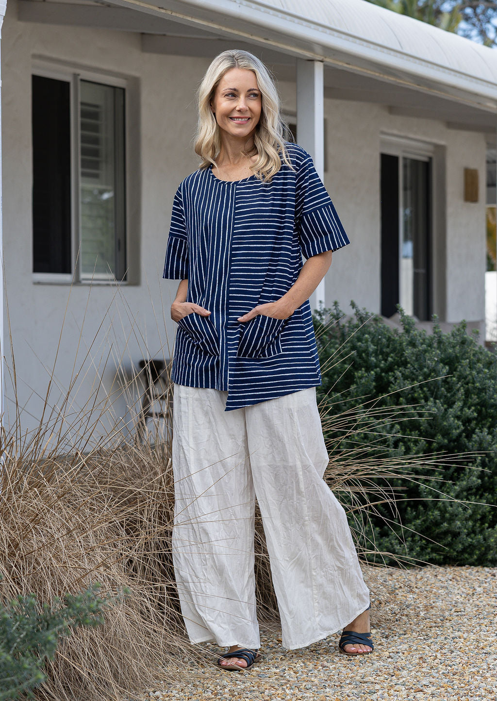 Woman wears a navy and white striped 100% cotton relaxed fit top with an asymmetrical hem line and front pockets paired with white pants