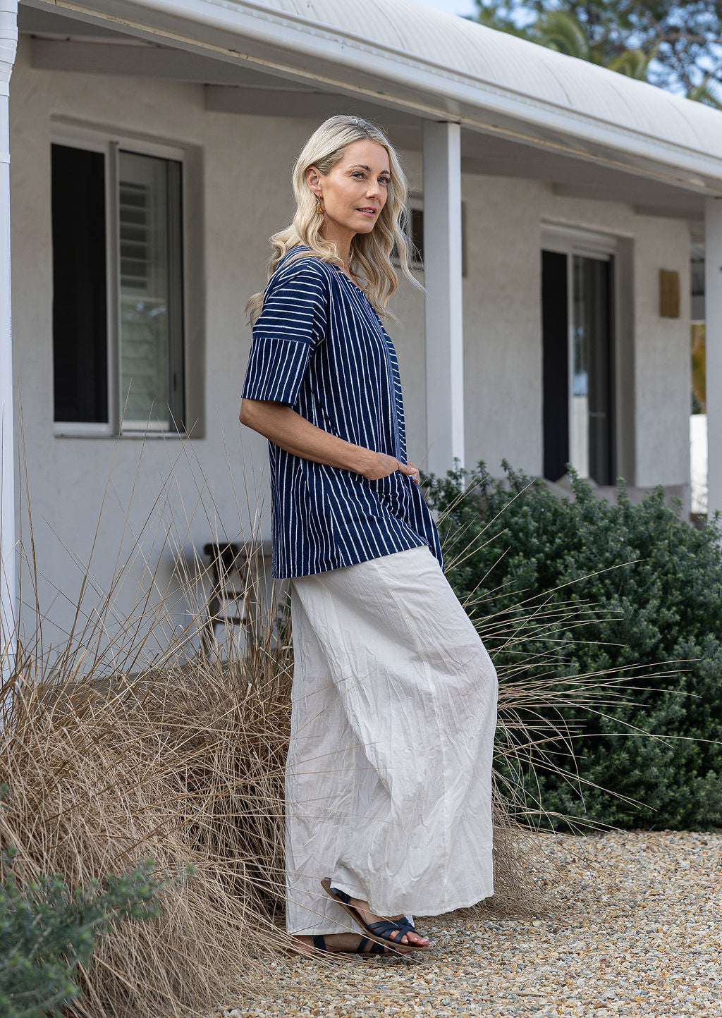 Woman standing sideways wearing a 100% cotton top in navy and white striped with both hand in pockets paired with white plain pants