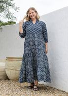 Woman wearing a 100% cotton tiered maxi dress in navy blue based floral print against a white wall 