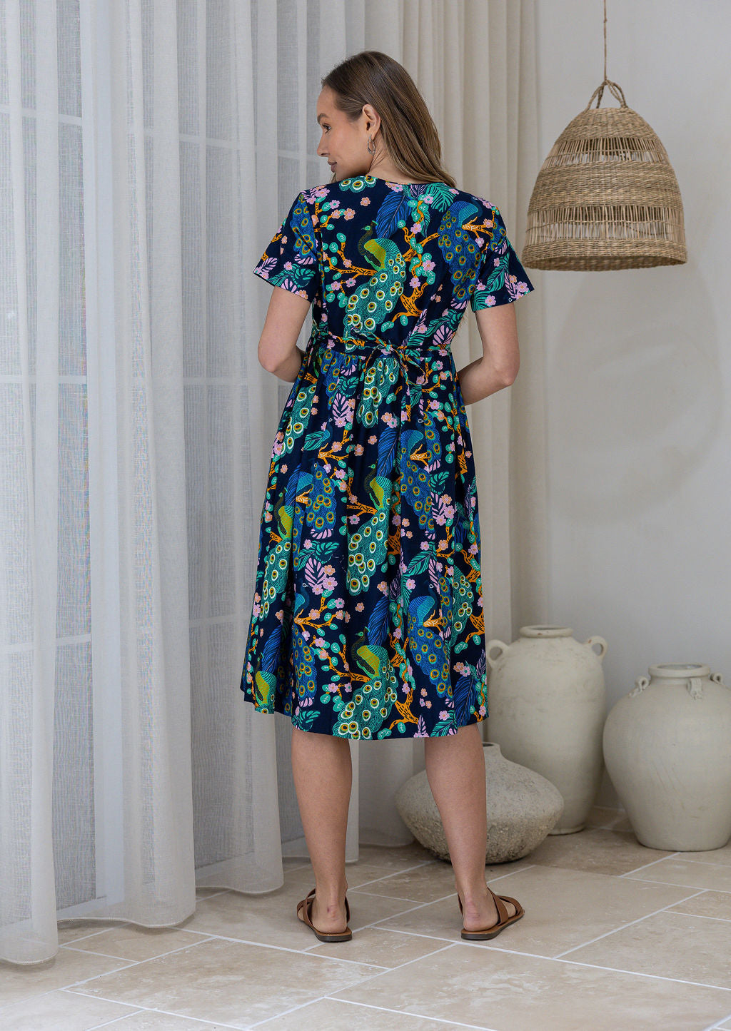 Woman in a navy based cotton floral dress standing in a room showing the back of the dress cinched with shoestring ties