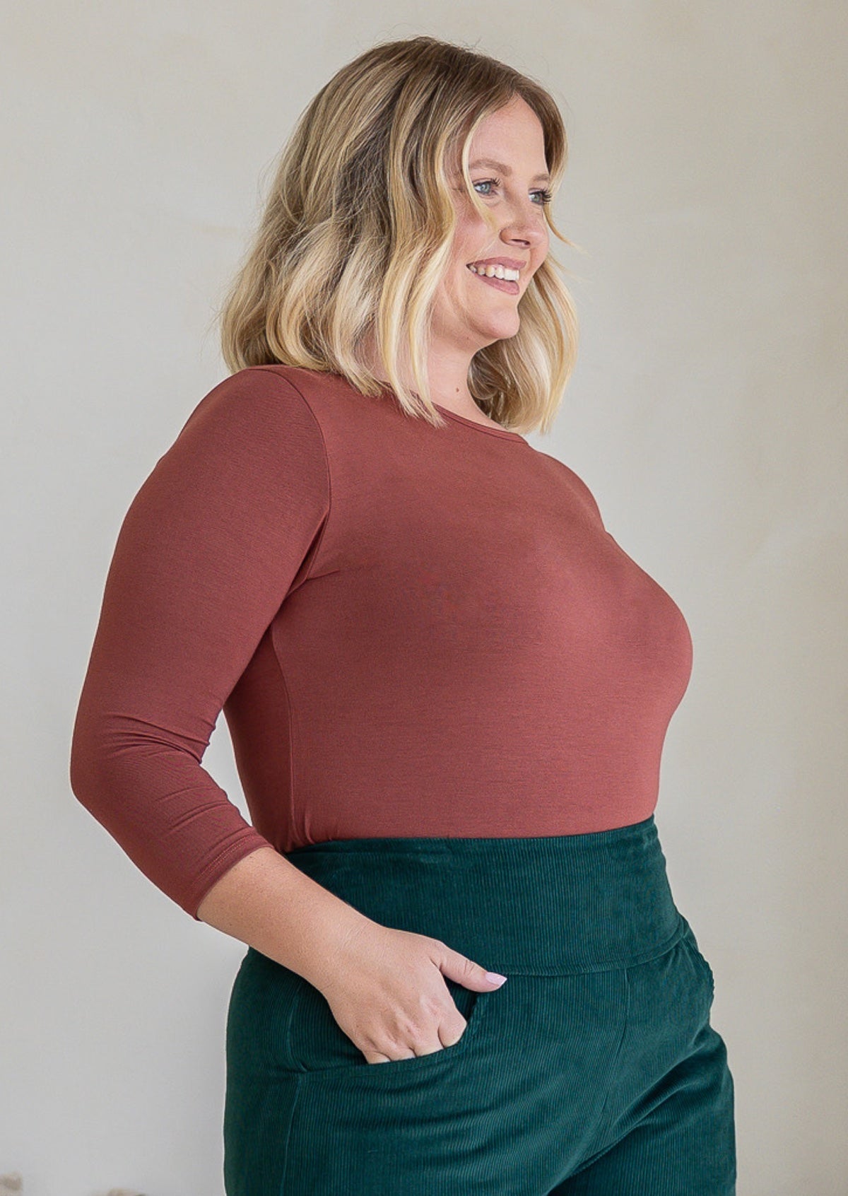 Woman standing sideways wearing a rayon boat neck terracotta pink 3/4 sleeve top.