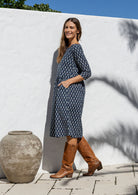 Model standing sideways wearing a relaxed fit cotton dress sitting just below the knee length with wide round neckline and hand in pockets.