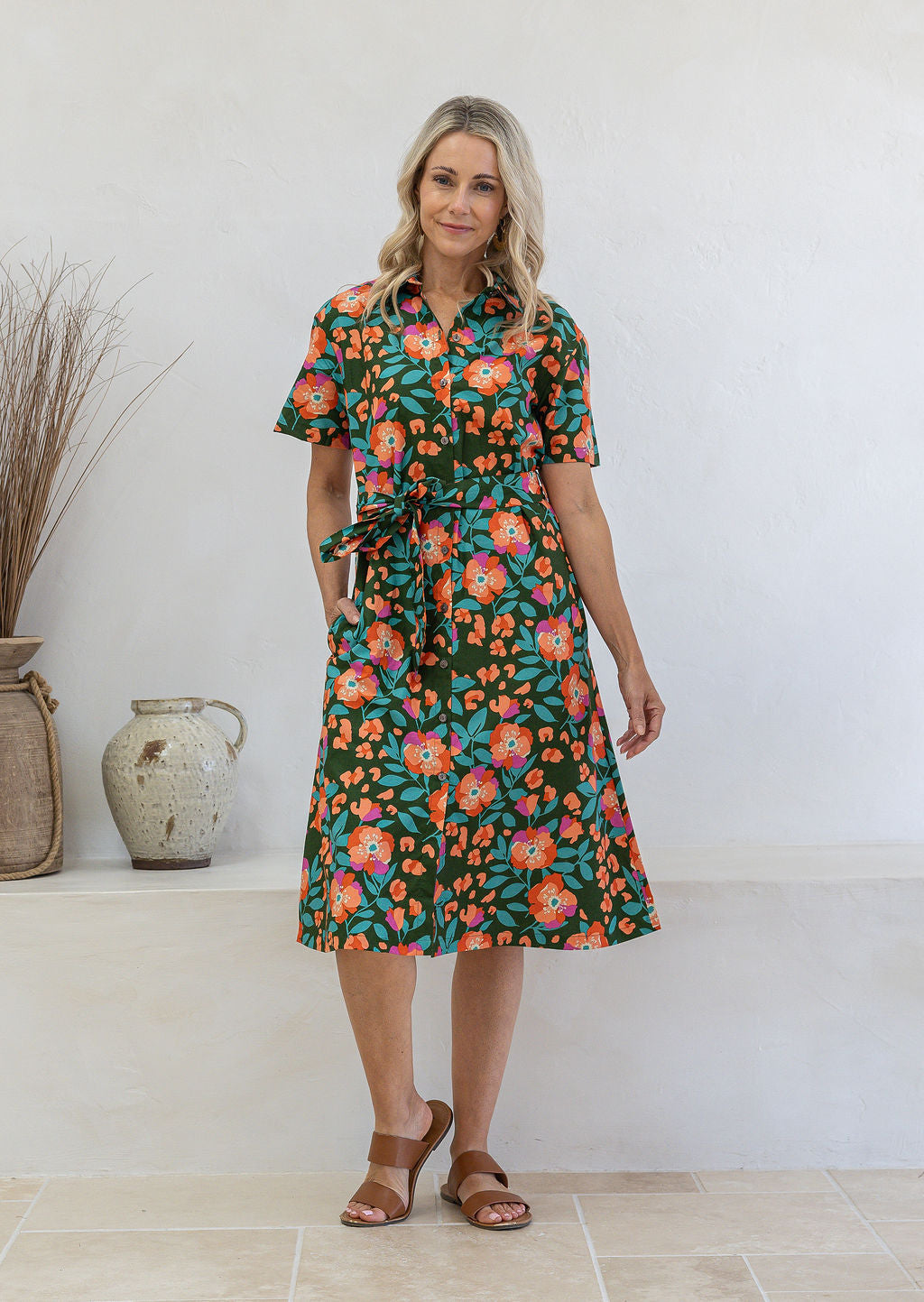Woman wearing 100 percent cotton button through shirt dress with collar and short sleeves, in a olive green based floral print.