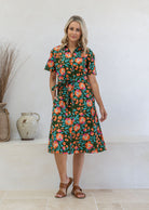 Woman wearing 100 percent cotton button through shirt dress with collar and short sleeves, in a olive green based floral print.