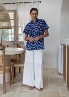 A woman stands in a bright, modern kitchen wearing a navy blue cotton short-sleeve relaxed fit button-up shirt with a white leaf pattern and white wide-leg pants.