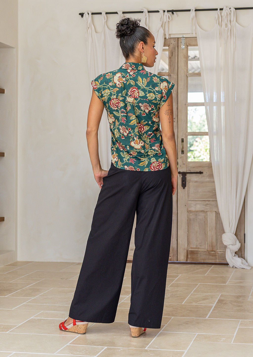 woman standing backwards wearing a green based floral printed slim fit shirt, in a room setting.