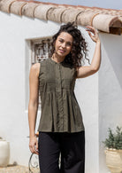 Woman wearing a lightweight 100% cotton olive green shade sleeveless top with pin tuck detailing and mandarin collar, standing in front of a rustic wooden window.