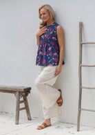 Woman wearing a floral sleeveless top with decorative pleats, high round neckline and white pants leaning against a white wall.
