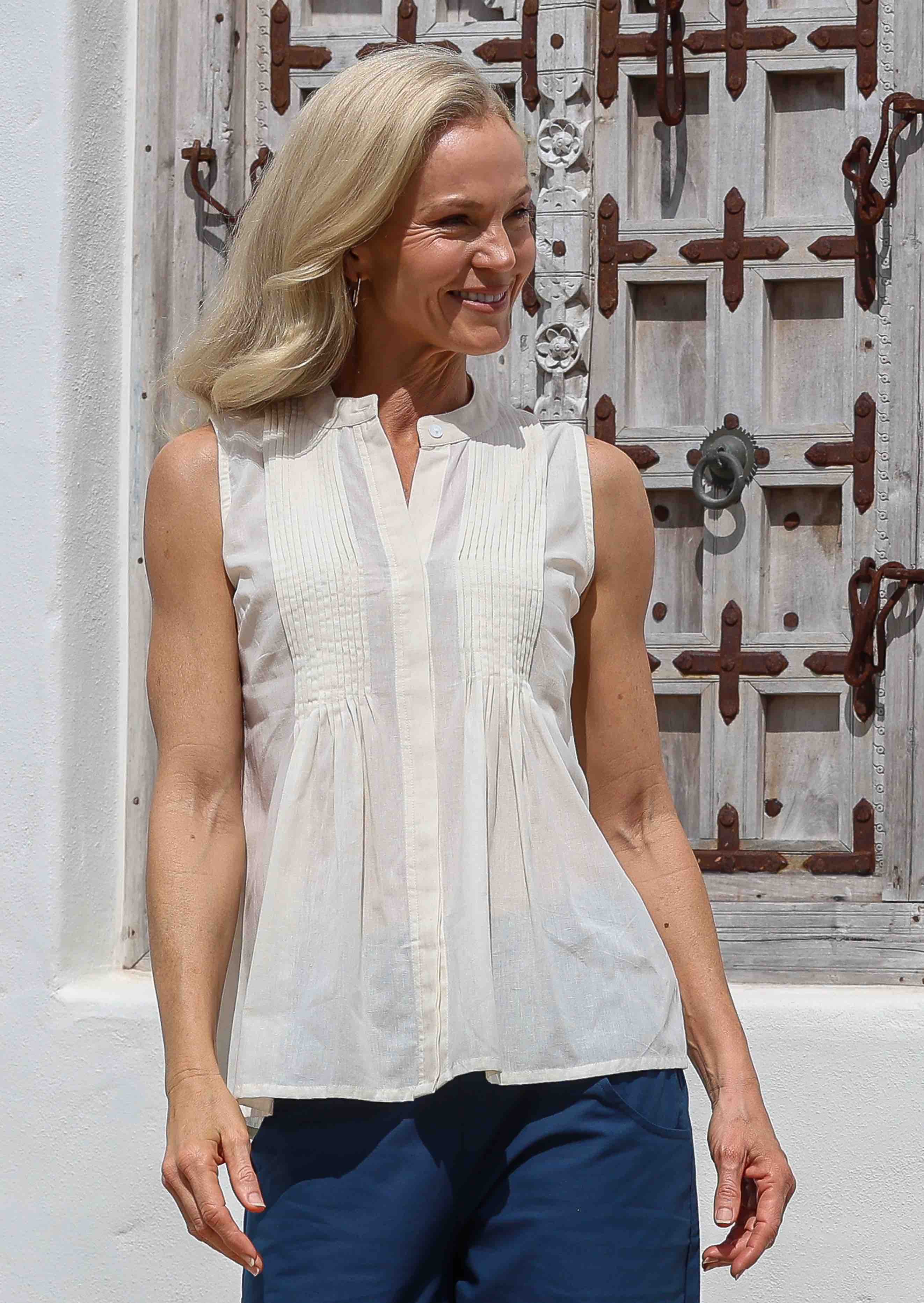 Woman wearing a white sleeveless cotton blouse with tiny pleats in front and blue pants standing in front of a decorative wooden window.