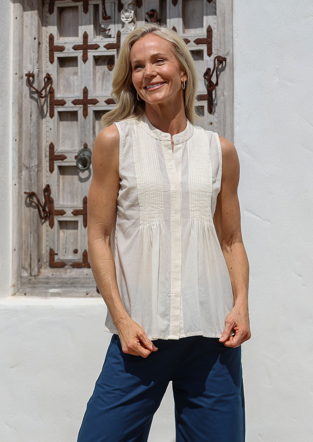 Woman wearing a lightweight 100% cotton white sleeveless top with pin tuck detailing and mandarin collar, standing in front of a rustic wooden window.